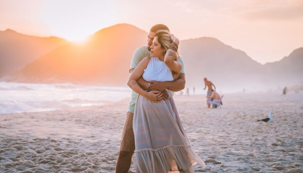 pregnant couple on a beach hugging